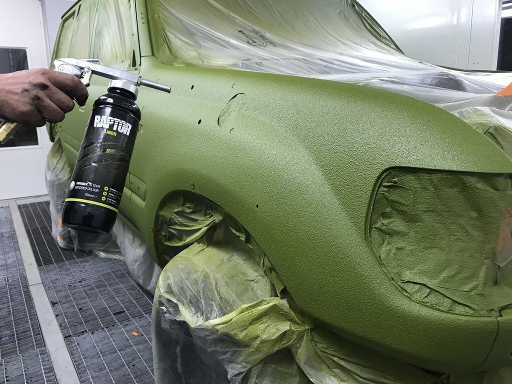 Hand spray painting a car with green Raptor Liner under protective covering in a workshop.