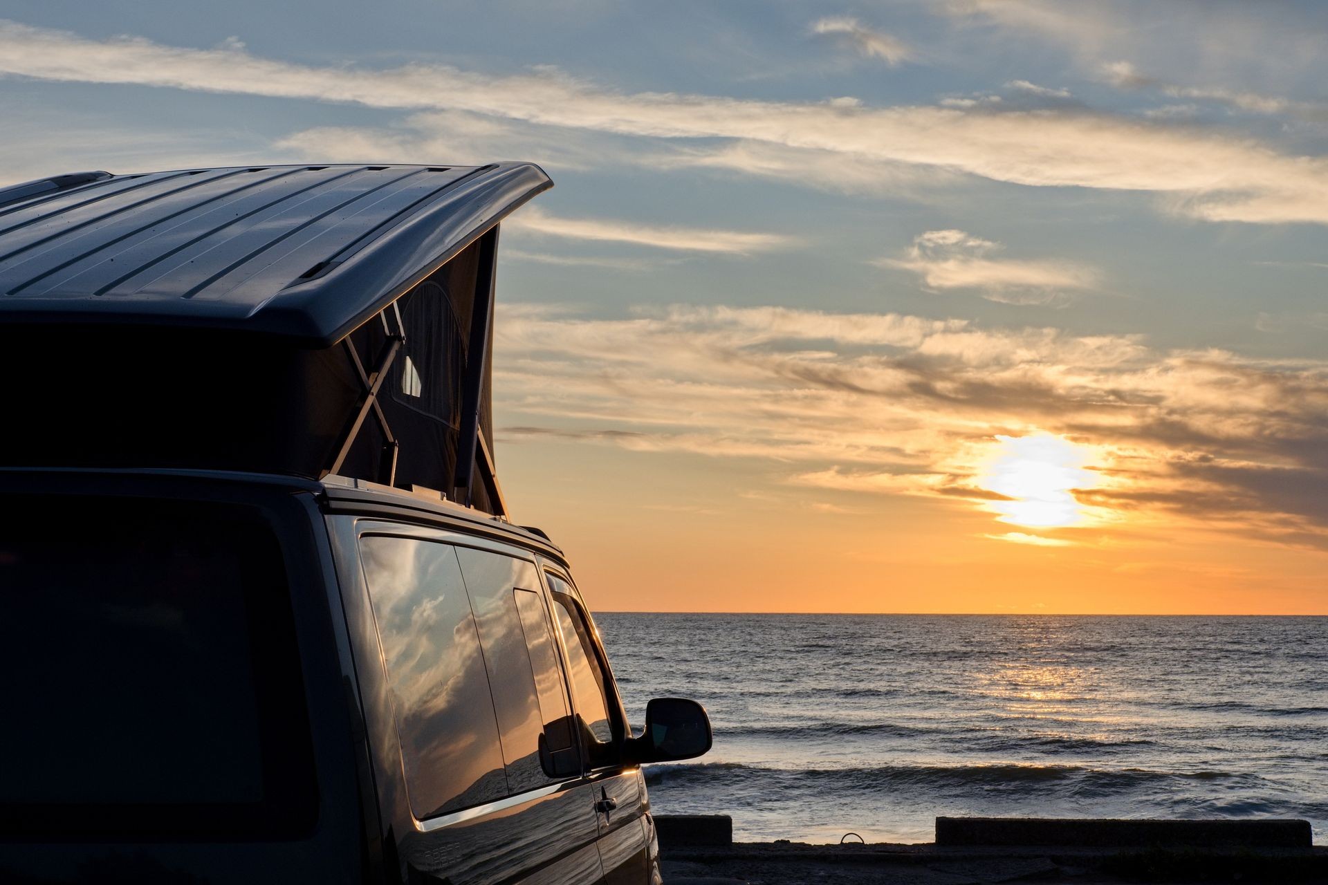 Camper van with open roof parked by the sea during sunset, reflecting light on its windows.